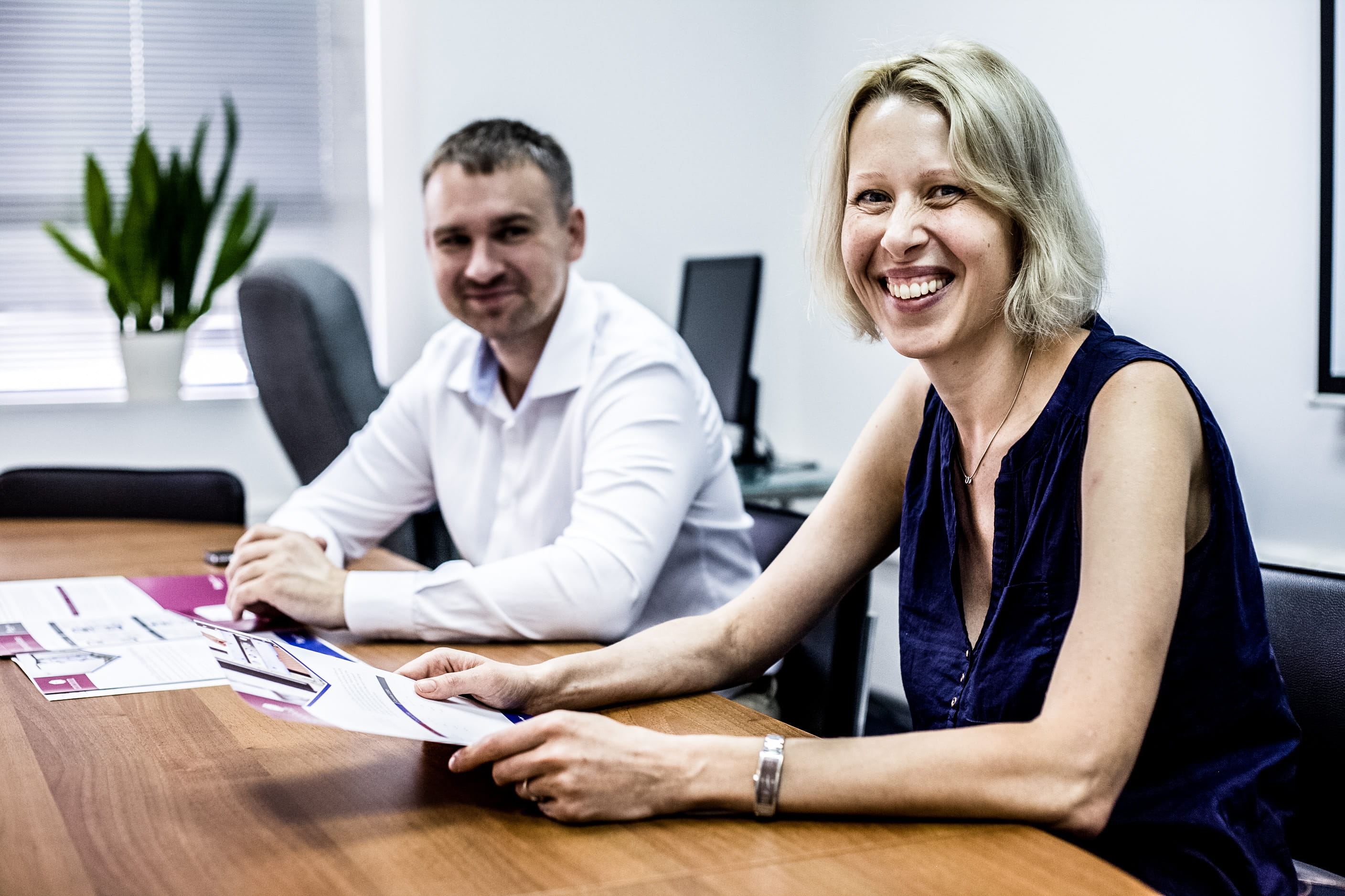 Two associates sitting at a table reviewing documents, look up and smile at the camera.