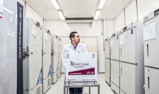 A man pushes a box on a cart down a wide aisle of storage lockers in a pharmaceutical storage depot.