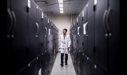 A man in a lab coat walks down a narrow aisle of lockers in a storage depot.