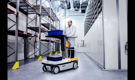 A man drives a forklift holding a small bucket down a wide aisle of shelves in a storage depot.
